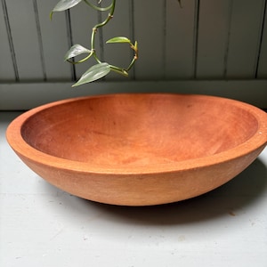 May include: A round, shallow wooden bowl with a warm, reddish-brown tone and a visible natural grain texture. The bowl features a smooth, curved interior and a thick, rounded rim, presented as a standalone object with a simple, functional design.