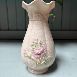 May include: A pale pink ceramic vase with a textured, pebbled surface and a ruffled rim. The body features a raised, molded floral design in soft pink and green tones, with a decorative band of scalloped detailing just below the neck.