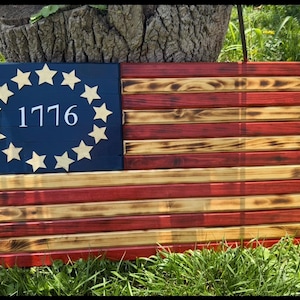 Peut inclure: Une décoration murale rectangulaire en bois en forme de drapeau avec des bandes horizontales rouges et en bois naturel et un canton bleu foncé. La section bleue présente treize étoiles claires disposées en cercle autour du texte blanc 1776.