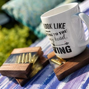May include: A white ceramic mug with black text that reads I LOOK LIKE I'M LISTENING TO YOU BUT IN MY HEAD I'M SHOOTING, placed next to two square coasters made of dark wood and clear resin containing metallic bullet casings.
