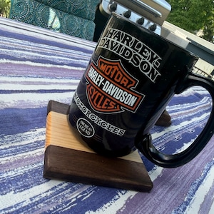 May include: A black ceramic mug featuring the Harley-Davidson logo and the text American Legend, resting on a square wooden coaster made of alternating light and dark wood strips. The mug has a large handle and a glossy finish with white and orange graphics.