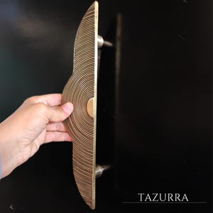 May include: A decorative brass door handle with a circular design. The handle is held against a black surface. The handle has a series of concentric lines and a circular accent. The word "TAZURRA" is printed in white.