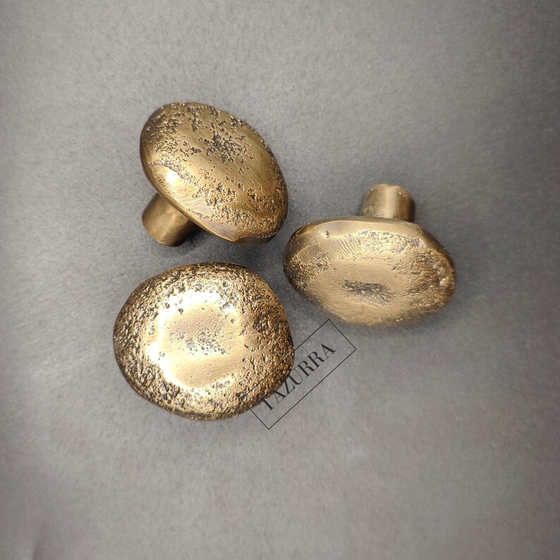 May include: Three brass mushroom-shaped cabinet knobs with a textured surface. The knobs are in a warm gold colour and are arranged on a grey background. The knobs are likely for furniture or decorative use.