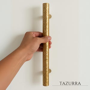 Könnte beinhalten: Ein goldfarbener, gehammerter Metall-Türgriff mit zylindrischer Form. Der Griff wird von einer Hand vor einem weißen Hintergrund gehalten. Das Wort "TAZURRA" ist unten rechts in schwarzer Schrift gedruckt.