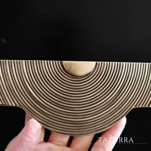 May include: A close-up of a brass-toned cabinet pull with a half-circle design of concentric lines. The pull has a rounded center and is held by a hand. The word "TAZURRA" is visible in the bottom right corner.