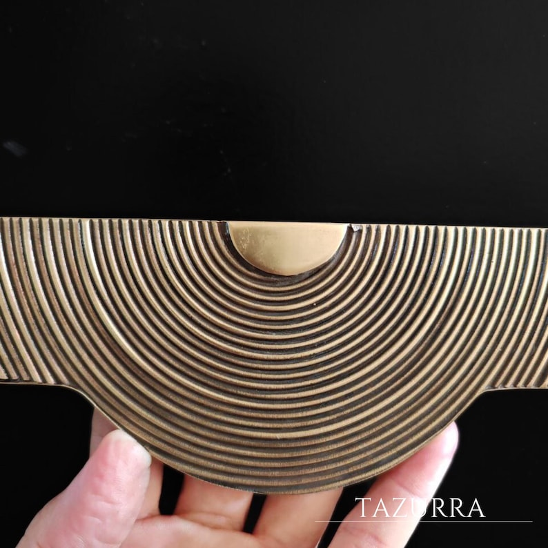 May include: A close-up of a brass-toned cabinet pull with a half-circle design of concentric lines. The pull has a rounded center and is held by a hand. The word "TAZURRA" is visible in the bottom right corner.