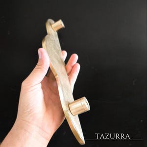May include: A brass door handle with a unique, elongated shape and two cylindrical mounting points. The handle has a warm, golden hue and a textured surface. The word "TAZURRA" is printed on the bottom right corner.