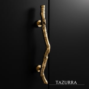 Peut inclure: Une poignée de porte dorée en forme de branche avec une surface texturée. La poignée est montée sur une porte noire. Le mot "TAZURRA" est imprimé en blanc en bas à droite.