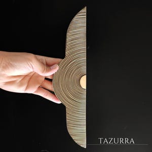 May include: A brass-toned door pull with a half-circle design featuring concentric circles and parallel lines. The text "TAZURRA" is visible in the lower right corner against a black background. The door pull is held by a hand.