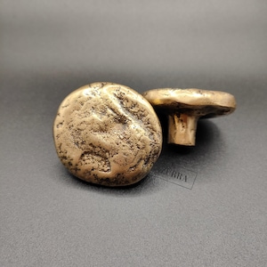 May include: Two antique-style cabinet knobs. The knobs have a textured, weathered appearance and are a warm, metallic gold colour. One knob is in focus, while the other is slightly out of focus. The knobs are a rounded, irregular shape.