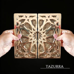 May include: Two rectangular, gold-colored metal panels with an intricate geometric design. The panels are held up against a black background. The word "TAZURRA" is printed in white at the bottom of the image.