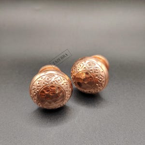 Könnte beinhalten: Zwei kupferfarbene Schrankknöpfe mit aufwendigen, geprägten Mustern. Die Knöpfe haben eine runde Form mit einer gehämmerten Textur auf der Vorderseite und einem dekorativen Design an den Rändern. Das Wort "TAZURRA" ist auf dem Bild aufgedruckt.