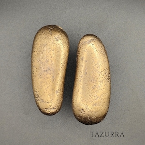 Puede incluir: Dos objetos oblongos de color dorado con una superficie texturizada se muestran sobre un fondo gris. Los objetos tienen forma redondeada y parecen ser de metal. La palabra "TAZURRA" está impresa en la parte inferior.