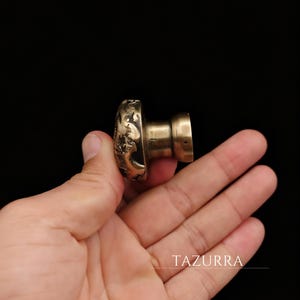 Könnte beinhalten: Ein bronzefarbener, pilzförmiger Türknauf mit aufwendigen Reliefschnitzereien. Der Knauf hat einen zylindrischen Schaft und einen abgerundeten, dekorativen Kopf. Das Bild ist vor schwarzem Hintergrund.