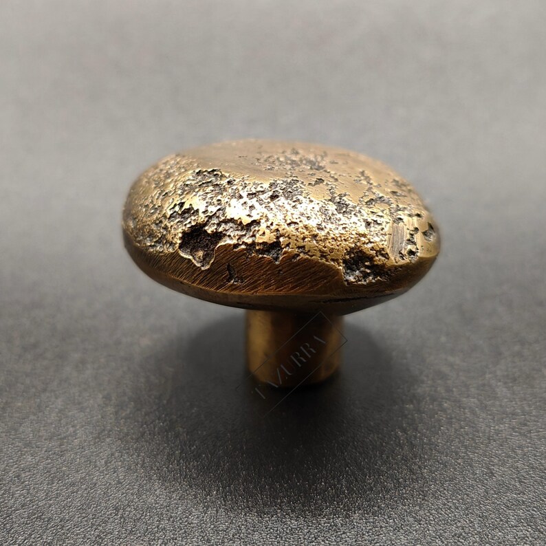 May include: A bronze cabinet knob with a textured, pitted surface. The knob has a rounded, mushroom-like shape and a short, cylindrical stem. The finish is a warm, antique gold colour, suggesting a vintage or rustic aesthetic.