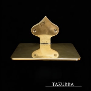 May include: A gold-colored, wall-mounted shelf with a decorative, spade-shaped top. The shelf has a rectangular base and is attached to the wall with two screws. The word "TAZURRA" is printed at the bottom of the image.