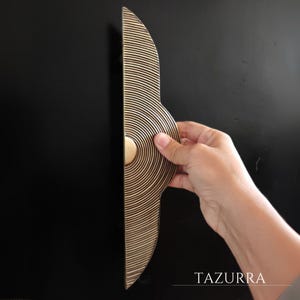 May include: A decorative door handle with a unique design. The handle is made of a light brown material with concentric circular grooves and a brass-colored circular accent. The handle is held against a black surface. The word "TAZURRA" is in the image.