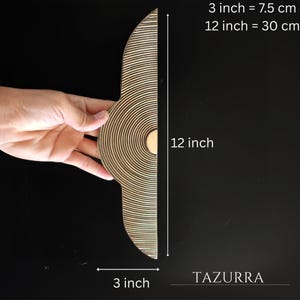 May include: A wooden decorative piece with a unique design. The piece is 12 inches (30 cm) long and 3 inches (7.5 cm) wide. The design features concentric circles on one side and parallel lines on the other. The brand name "TAZURRA" is visible.