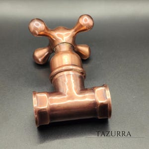 May include: A copper-colored faucet with a cross-shaped handle. The faucet has a T-shaped body with threaded ends. The word "TAZURRA" is printed at the bottom right corner. The faucet is made of metal and has a shiny finish.