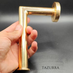 May include: A brass-colored, L-shaped door pull or handle. The handle is held in a hand against a dark background. The text "TAZURRA" is visible at the bottom right.