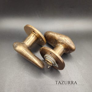 Puede incluir: Dos manijas de puerta antiguas de bronce con un acabado envejecido. Cada manija tiene una base redonda y una palanca curva. Las manijas se muestran sobre una superficie oscura. La palabra "TAZURRA" está impresa debajo.