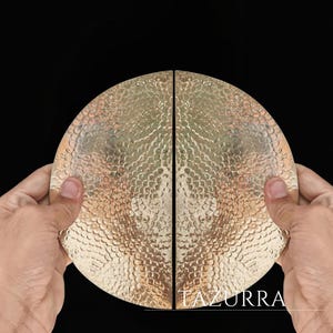 May include: Two halves of a gold-coloured, hammered metal plate, held together. The plates have a textured surface with a pattern of small, evenly spaced indentations. The word "LAZURRA" is printed in white.