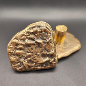 May include: A bronze-colored, abstract sculpture with a textured, rock-like form. A cylindrical piece is attached to a flat, organic base. The sculpture has a weathered appearance, suggesting age or a natural aesthetic.