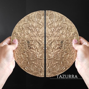 May include: Two gold-coloured, half-circle door pulls with a textured surface. The door pulls are held by hands against a black background. The word "TAZURRA" is printed at the bottom.