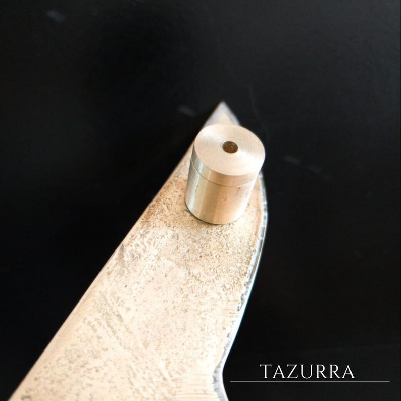 May include: A cylindrical, brass-colored object with a hole in the top rests on a textured, metallic surface. The surface is shaped like a blade. The word "TAZURRA" is printed in white on the lower right corner.