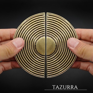 May include: Two antique brass-colored cabinet pulls, each a half-circle with concentric ridges. The pulls are held together, forming a full circle. The word "TAZURRA" is printed at the bottom.