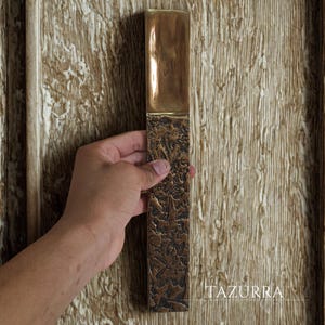 May include: A rectangular bronze door pull with a textured lower section and a smooth, polished upper section. The door pull is held against a wooden door. The word "TAZURRA" is printed on the door.