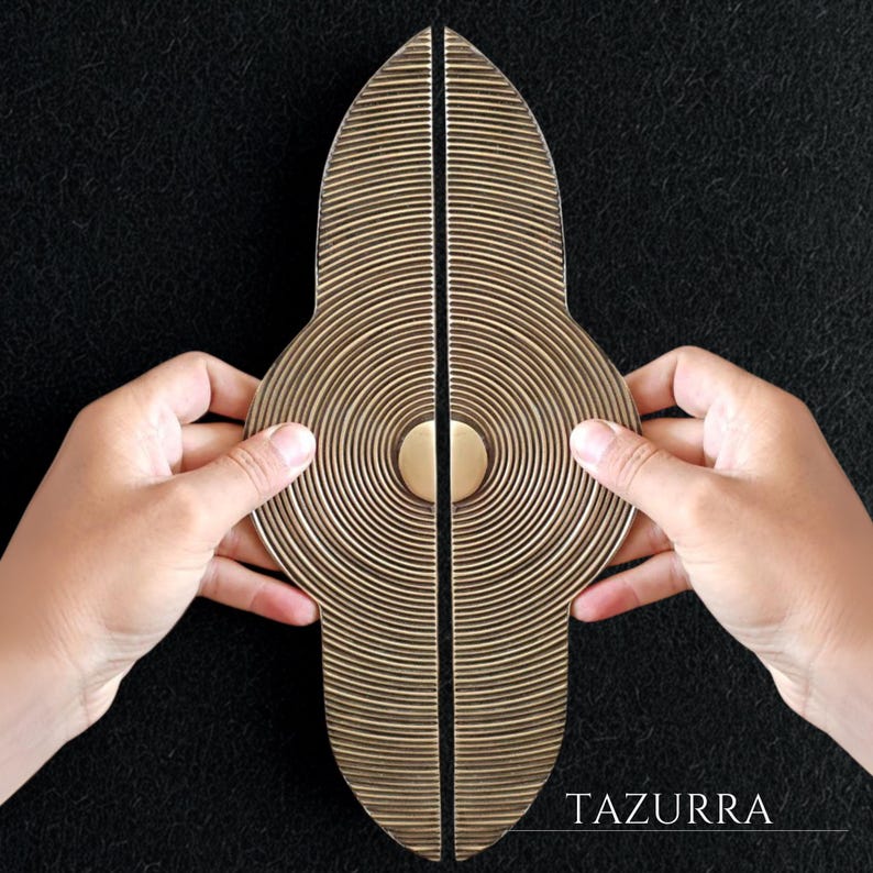 May include: Two brass-colored door handles with a unique design. Each handle is a half-oval shape with concentric circular grooves and a central circular accent. The handles are held together, with the word "TAZURRA" visible in the lower right corner.