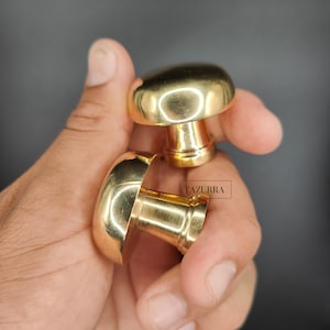 May include: Two shiny, gold-colored mushroom-shaped cabinet knobs. The knobs have a rounded top and a cylindrical base. The word "TAZURRA" is printed on the base of one of the knobs. These knobs are likely used for furniture or decorative purposes.
