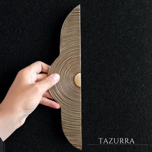 Peut inclure: Une poignée de porte en laiton en forme de demi-lune avec des rainures circulaires concentriques. La poignée a un petit accent rond de couleur claire. Le mot "TAZURRA" est imprimé en blanc en bas à droite.