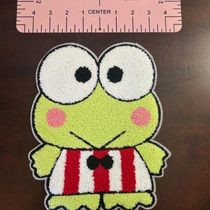 May include: A green frog-like character patch with large white eyes, black pupils, and pink cheeks. The frog wears a red and white striped shirt with a black bow tie. A pink V-neck back guide is at the top.