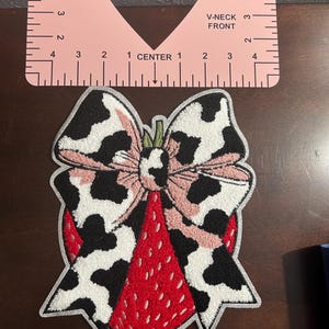 May include: A decorative patch featuring a strawberry design with a bow. The bow has a cow print pattern with pink accents. The strawberry is red with a textured appearance, and the patch has a silver border. A pink V-neck guide is in the background.