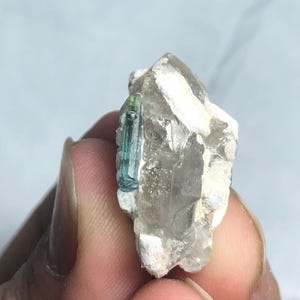 Könnte beinhalten: Eine rohe, natürliche Kristallformation mit einer klaren Quarzbasis und einem kleinen, leuchtend blau-grünen Turmalinkristall. Der Quarz hat eine raue, strukturierte Oberfläche, und der Turmalin zeigt eine prismatische Struktur.