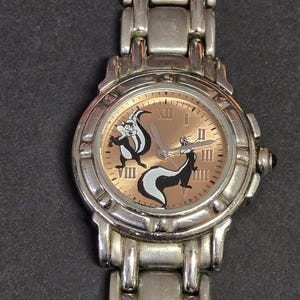 Pu&ograve; includere: Orologio da polso color argento con quadrante marrone chiaro, numeri romani e due personaggi dei cartoni animati di puzzole. L'orologio ha un cinturino argentato e un quadrante rotondo. Le puzzole sono nere e bianche.