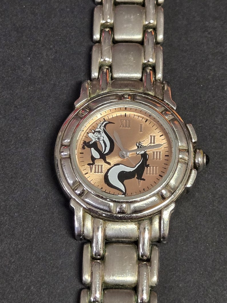 Pu&ograve; includere: Orologio da polso color argento con quadrante marrone chiaro, numeri romani e due personaggi dei cartoni animati di puzzole. L'orologio ha un cinturino argentato e un quadrante rotondo. Le puzzole sono nere e bianche.
