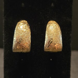 May include: A pair of gold-toned hoop earrings featuring a detailed, embossed floral pattern across the entire surface. The earrings have a semi-circular, thick design with a polished finish that catches the light, highlighting the intricate botanical engravings.