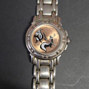 Pu&ograve; includere: Orologio da polso color argento con quadrante circolare con un design di puzzola dei cartoni animati. L'orologio ha un cinturino metallico con maglie rettangolari e un quadrante marrone chiaro. Le lancette dell'orologio sono nere.