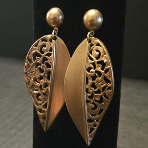 May include: A pair of gold-toned drop earrings featuring a leaf-shaped design. Each earring consists of a smooth, solid half and a detailed, openwork filigree half, suspended from a small, round, polished gold-toned stud post at the top.