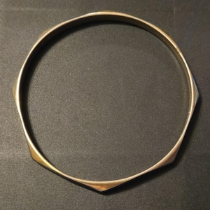 May include: A gold-toned, geometric bangle bracelet. The bracelet has a series of flat, angular facets that create a unique, modern design. The bracelet is circular and appears to be made of metal.
