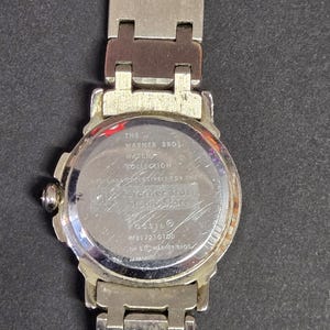 Pu&ograve; includere: Un orologio color argento della Warner Bros. Watch Collection. L'orologio ha un quadrante rotondo e un cinturino argentato. Il retro dell'orologio presenta la scritta "Warner Bros. Studio Store" e "Fossil".