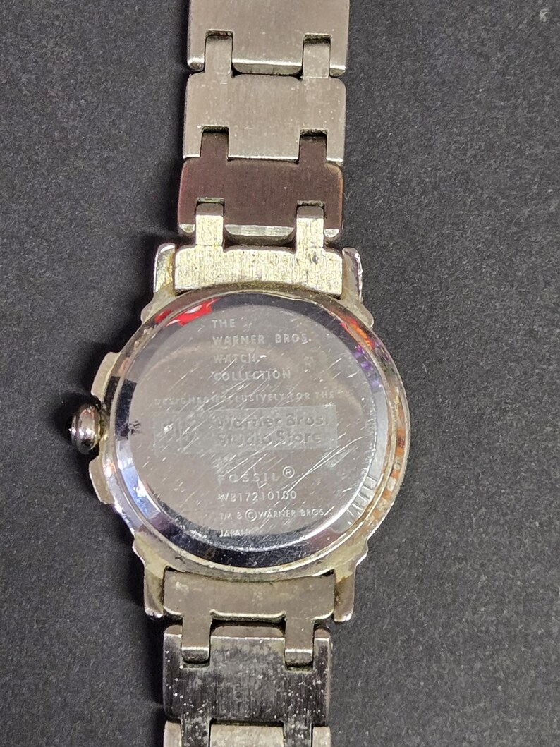 Pu&ograve; includere: Un orologio color argento della Warner Bros. Watch Collection. L'orologio ha un quadrante rotondo e un cinturino argentato. Il retro dell'orologio presenta la scritta "Warner Bros. Studio Store" e "Fossil".
