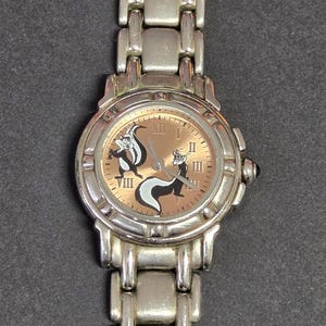 Pu&ograve; includere: Orologio da polso color argento con quadrante marrone chiaro, numeri romani e due moffette dei cartoni animati. L'orologio ha un cinturino argentato e una corona nera. Un orologio divertente e originale.