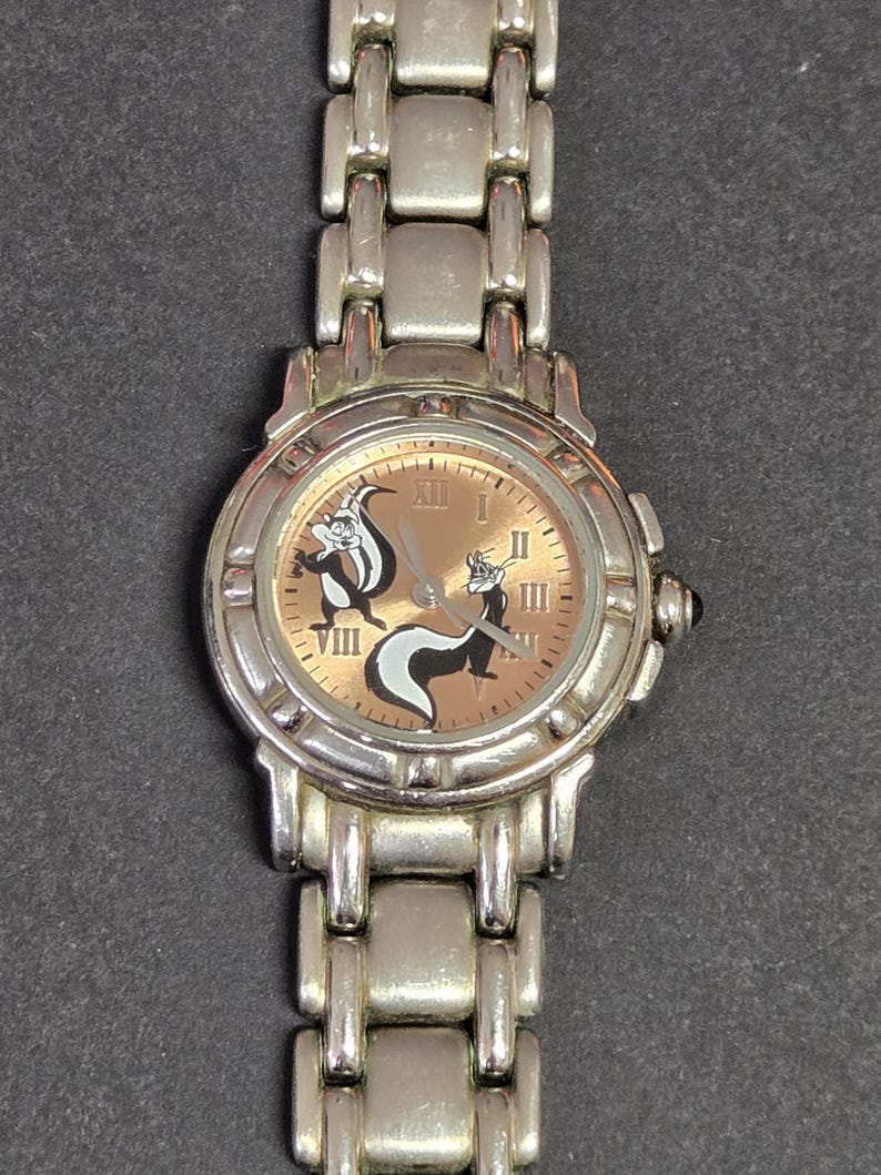 Pu&ograve; includere: Orologio da polso color argento con quadrante marrone chiaro, numeri romani e due moffette dei cartoni animati. L'orologio ha un cinturino argentato e una corona nera. Un orologio divertente e originale.