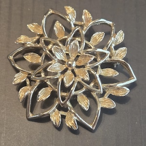 Könnte beinhalten: Goldfarbene Metallbrosche mit floralem Design. Die Brosche hat eine zentrale Blütenform, umgeben von durchbrochenen Blütenblättern und Blättern. Die Brosche ist kreisförmig und hat einen Nadelverschluss auf der Rückseite.