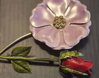 May include: A decorative floral brooch featuring a large, light purple flower with a gold center and a red rose. The brooch has a green stem with green leaves. The brooch is made of metal.
