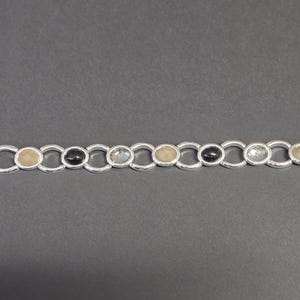 May include: Silver-toned bracelet featuring a series of oval-shaped gemstones in various colors, including black, clear, and beige. The bracelet has a toggle clasp closure. The gemstones are set within a chain-link design.